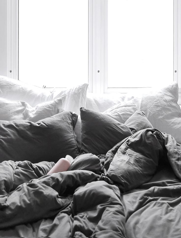 an unmade bed with pillows and blankets in front of two windows, one person laying on the floor