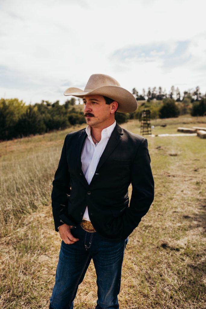 a man wearing a cowboy hat standing in a field with his hands on his hips