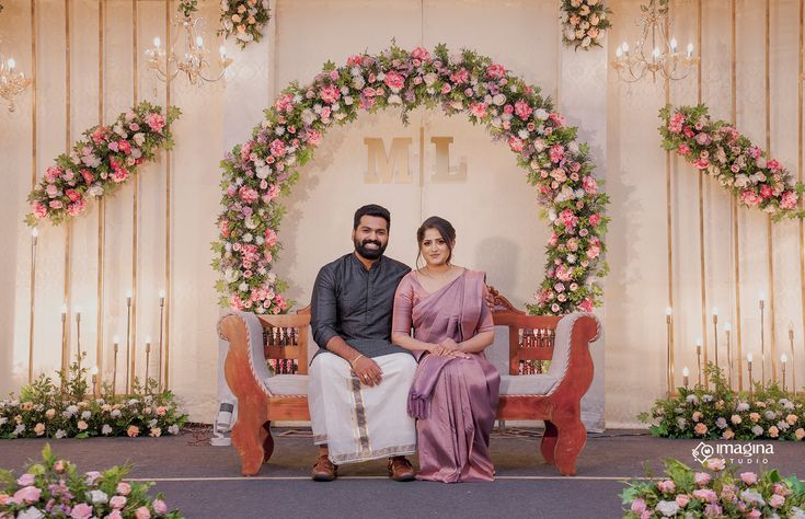 a man and woman sitting on a bench in front of a floral arch with flowers