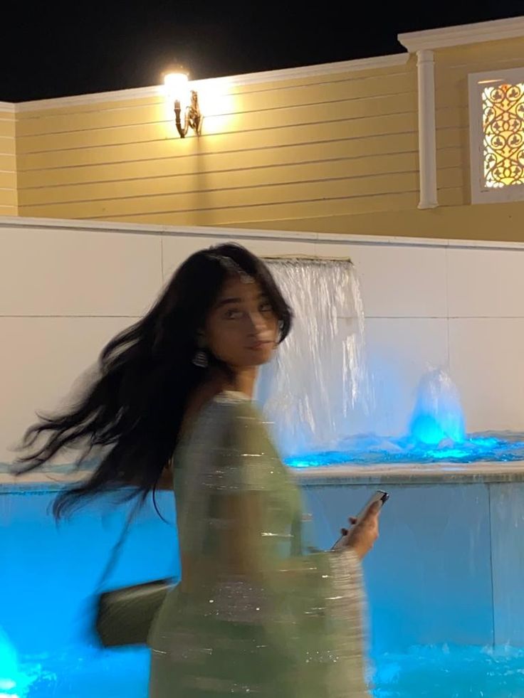 a woman is walking in front of a fountain at night with her cell phone up to her ear