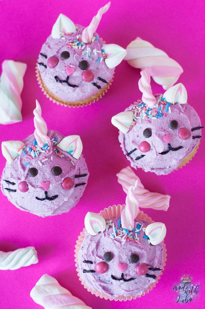 three cupcakes with pink frosting and cat ears on them, sitting next to marshmallows