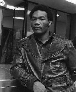 a man in a leather jacket holding a piece of paper and standing next to a desk