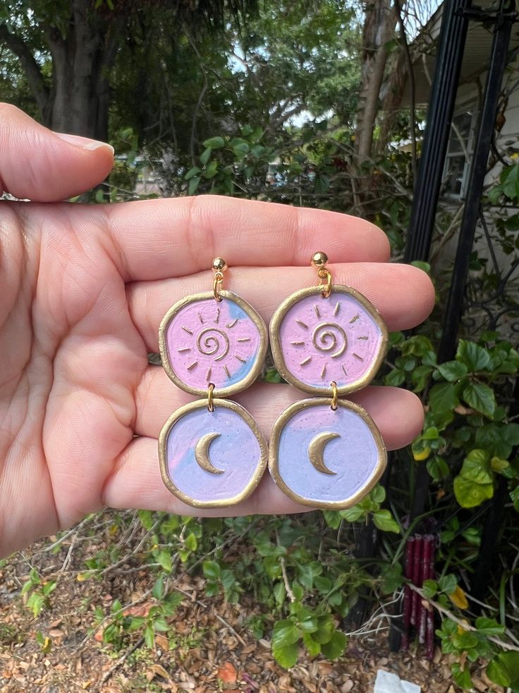 a pair of pink and purple earrings with crescents on them in front of some bushes