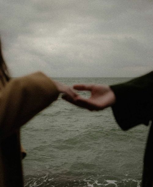 two people holding hands near the ocean on a gloomy day with waves coming in