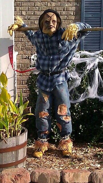 a statue of a scarecrow is posed in front of a brick wall and shrubbery