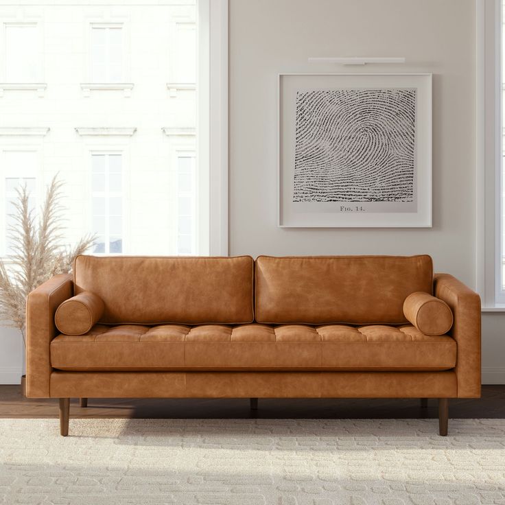 a tan leather couch sitting in front of a white wall with windows and a rug on the floor