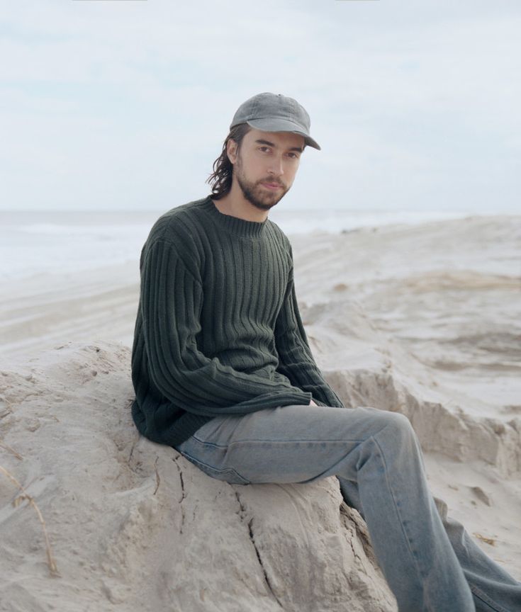 a man sitting on top of a rock next to the ocean wearing a baseball cap