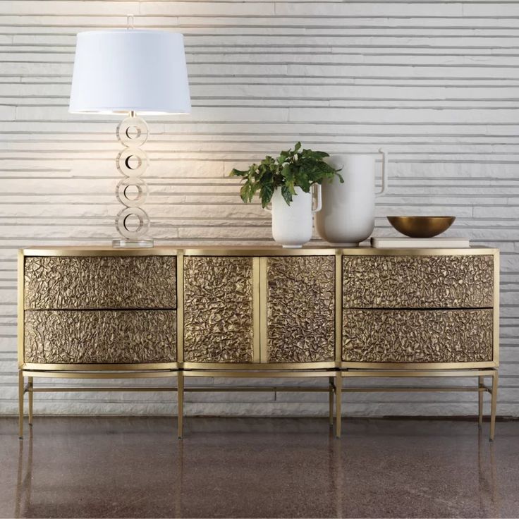 a gold sideboard with two white vases and a plant on it next to a lamp