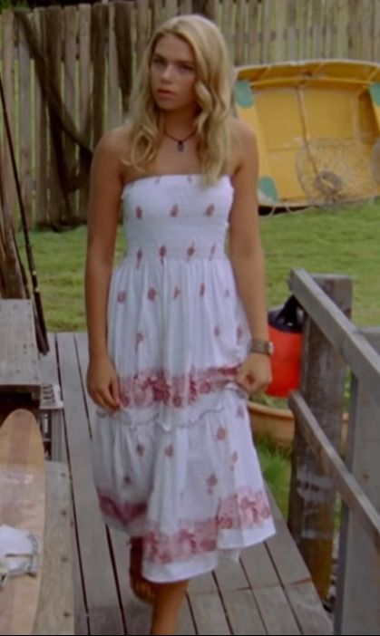 a woman in a white dress standing on a wooden deck next to a surfboard