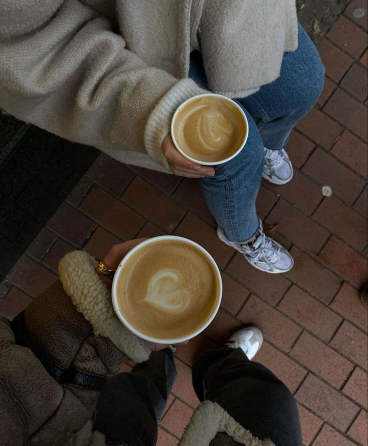 two people holding cups of coffee on top of a brick floor next to each other