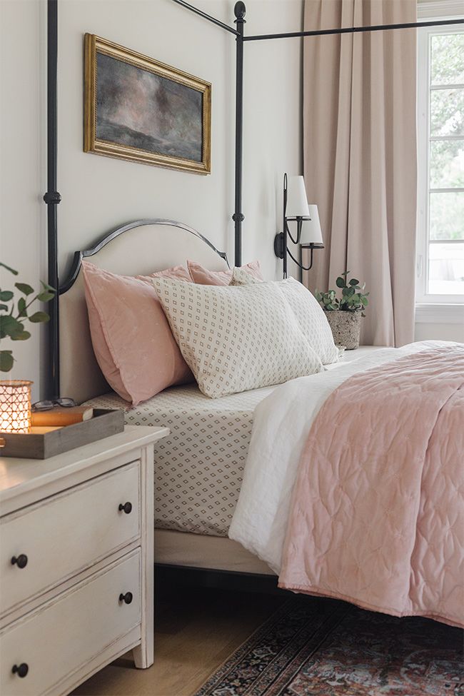 a bed with pink and white comforter in a bedroom next to a dresser, lamp and window