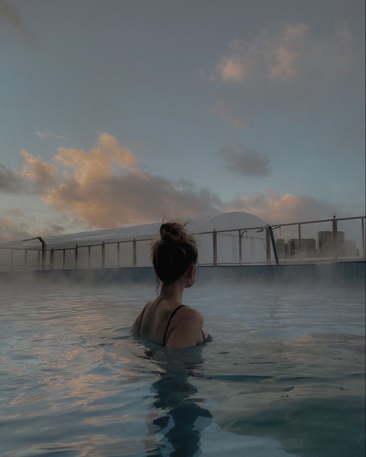 a woman in a swimming pool with the sun setting behind her and buildings on the horizon