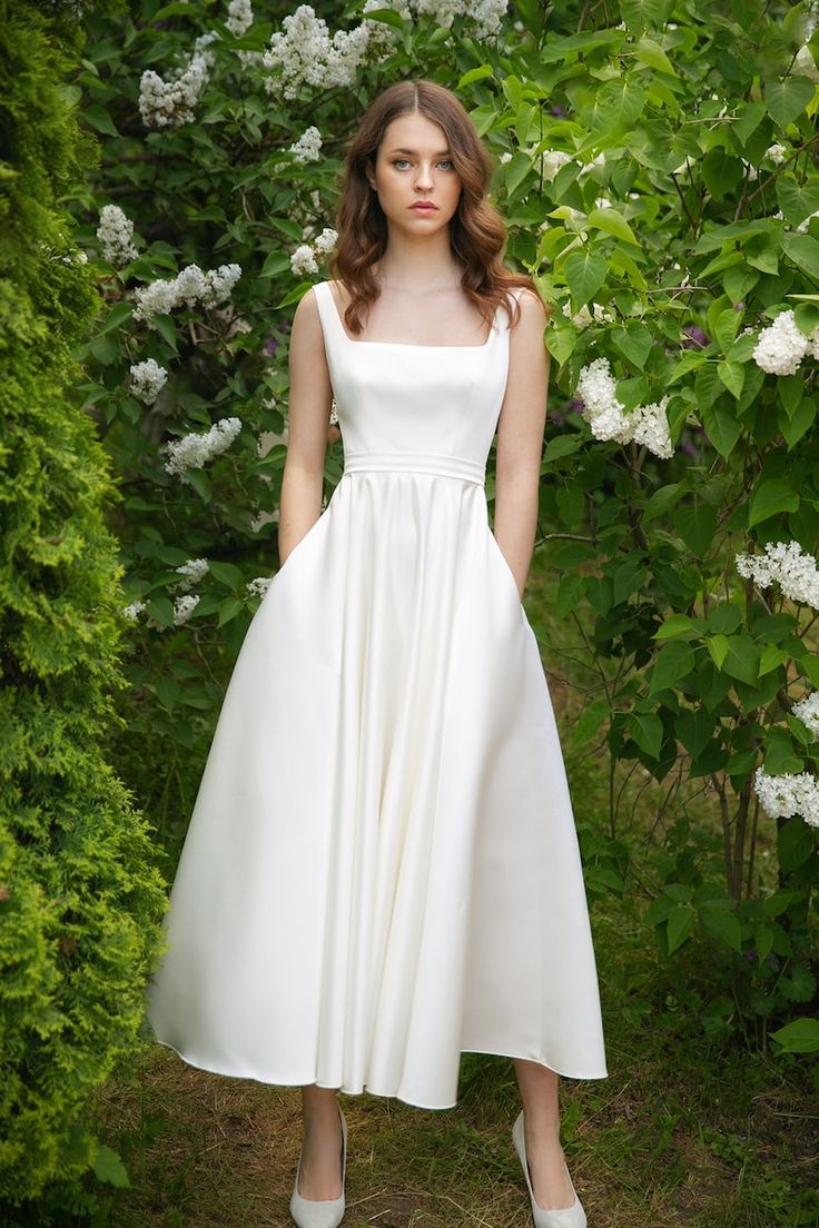 a woman in a white dress standing next to some bushes and flowers with her hands on her hips