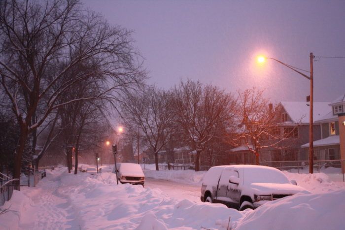 a snowy street with cars parked on the side and one car covered in snow at night