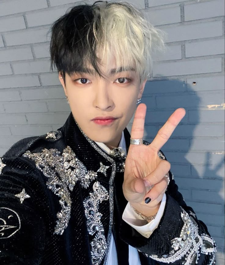 a man with white hair making the peace sign in front of a brick wall wearing a black and white jacket