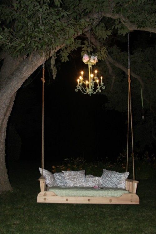 a wooden swing bed with pillows on it under a chandelier hanging from a tree