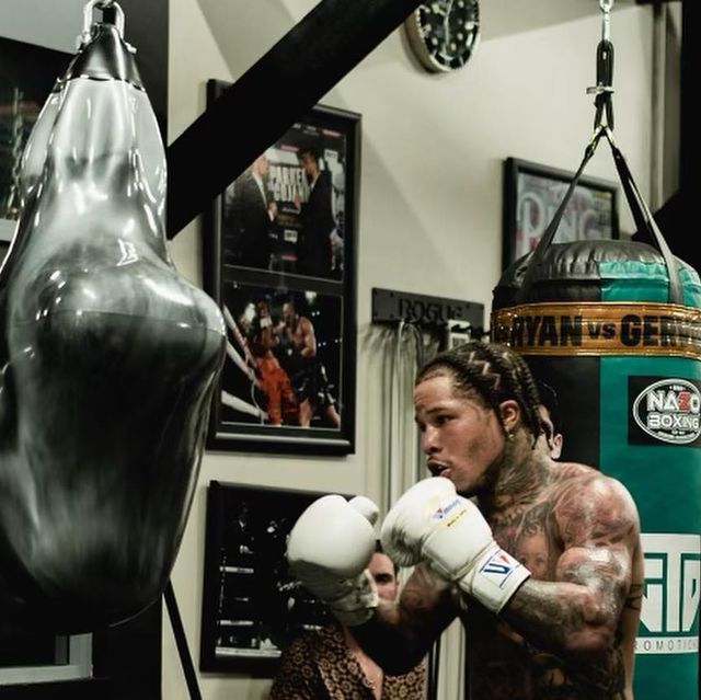 a man with long hair and tattoos wearing boxing gloves in a gym next to a punching bag