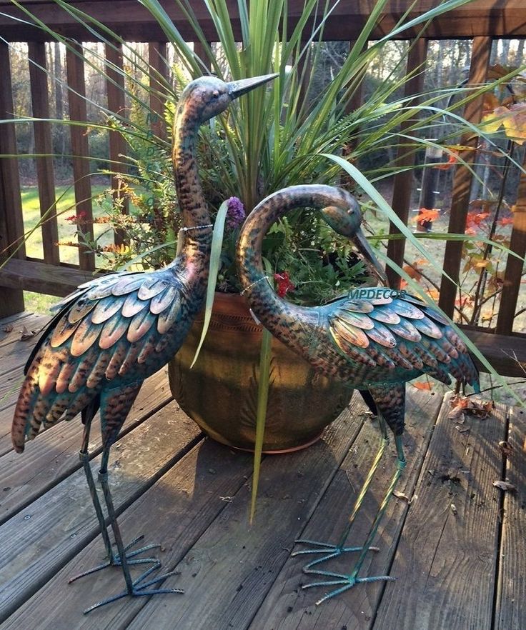 two metal birds are standing next to each other on a wooden deck with plants in it