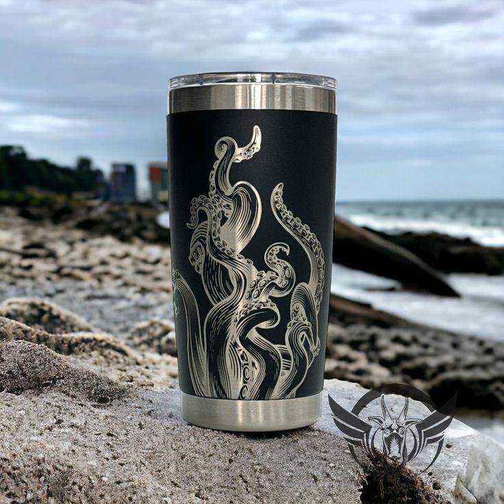 a black and silver cup sitting on top of a rock next to the ocean with an octopus drawn on it