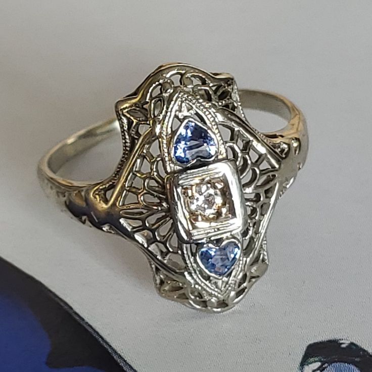 "Art Deco Navette Diamond Sapphire Filigree Ring. This piece is tested 18K white gold cast fashion lady's ring sizeable 7 1/2 measuring 18.35.. x 13.66mm with bright polish finish weighting 2.79 grams. The featured ring is Navette style and delicate filigree design rectangular round corners curved top with pierced out filigree and milgrain details centered by a square double bezel box holding a prong set round Old Mine cut Diamond measuring 2.55mm approx. 0.10ct guarded by each side with a pear Antique Hallmarked Cluster Ring For Anniversary, Fine Jewelry Sapphire Ring With Filigree, Heirloom Diamond Heart Ring For Wedding, Victorian White Gold Rings Stamped 14k, Antique Filigree Ring Stamped 14k For Anniversary, Collectible Yellow Gold Rings With Diamond Accents, Elegant White Gold Engraved Ring Stamped 14k, Formal Heart-shaped Jewelry With Intricate Design, Heart-shaped Jewelry With Intricate Design For Formal Occasions