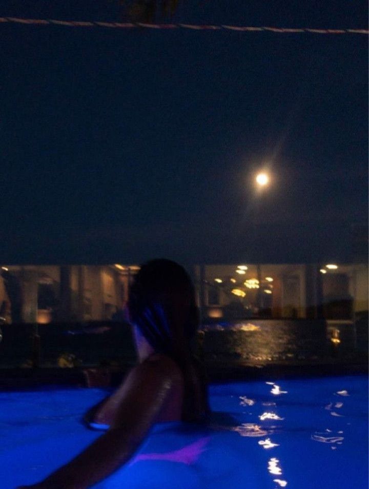 a woman sitting in a pool at night with the moon above her head and lights on