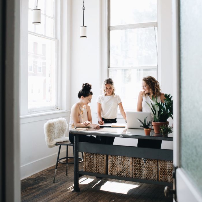 three women sitting at a table working on their laptops in front of a window