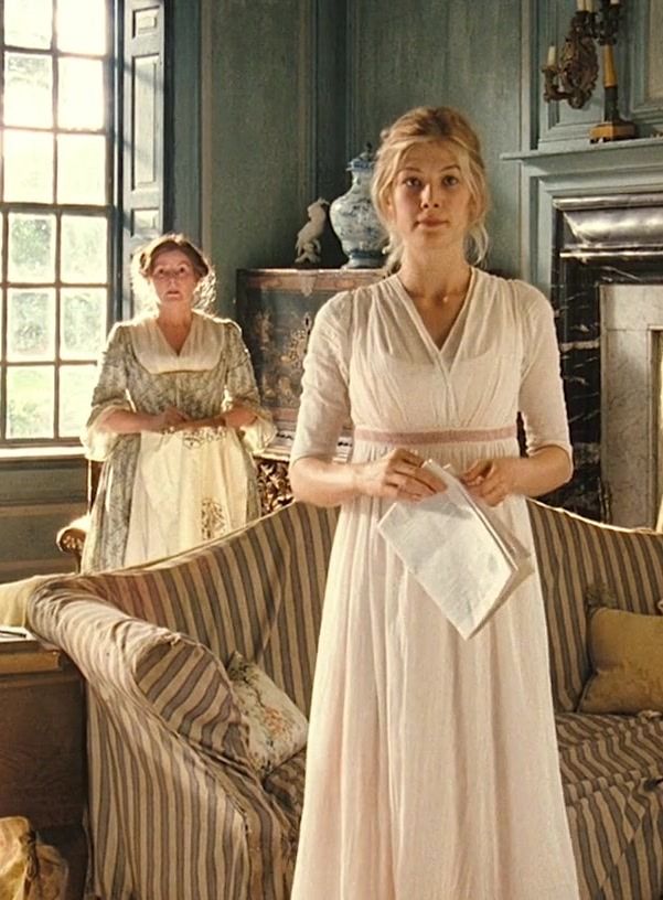 two women in period dress standing next to each other near a couch and fireplace with one woman looking at the camera