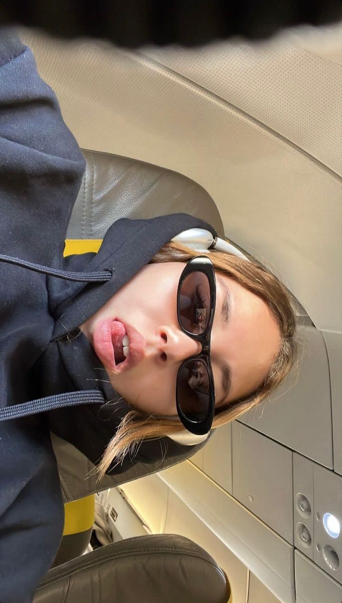 a woman wearing sunglasses and a hoodie sticking her tongue out while sitting on an airplane