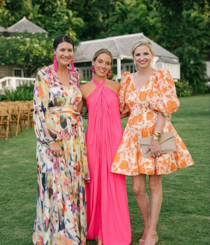 three women in dresses standing next to each other on the grass with trees in the background