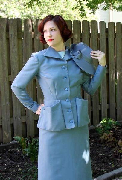 a woman in a blue dress and jacket standing next to a fence with her hands on her hips