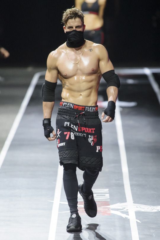 a shirtless man walking down a runway wearing black gloves and bandana on his face