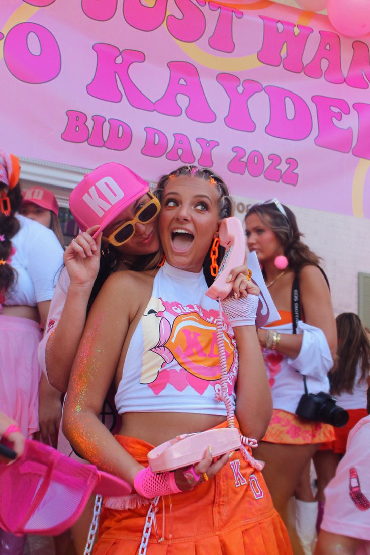 two women in pink and orange outfits are posing for the camera, with one holding a cell phone up to her ear