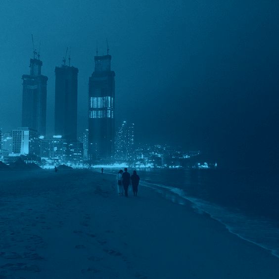 two people are walking on the beach in the dark with buildings in the background at night