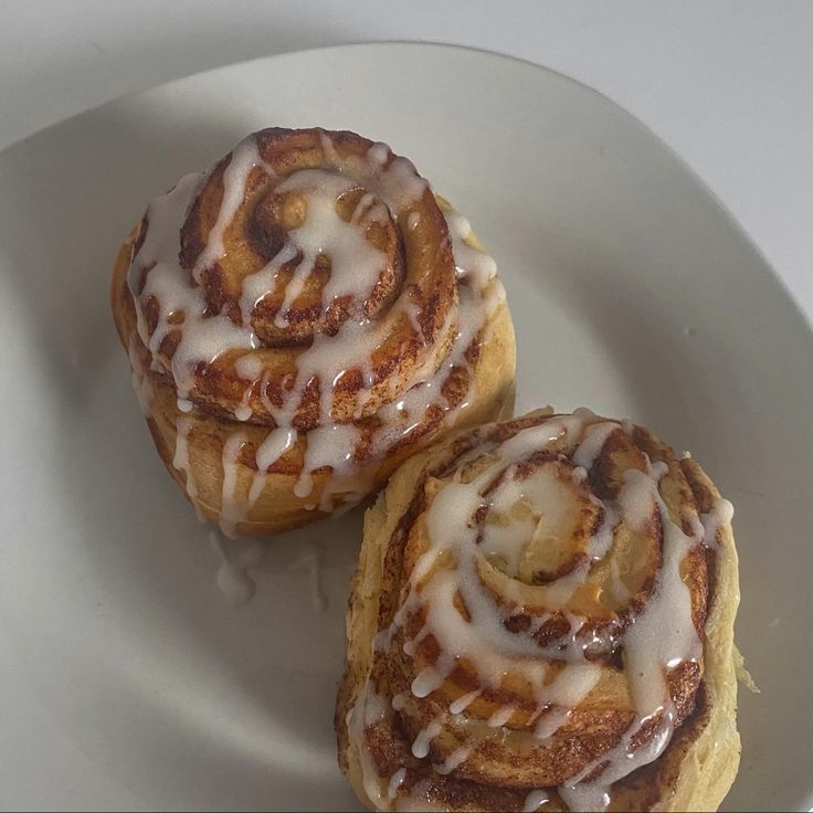 two cinnamon rolls on a white plate with icing drizzled around them