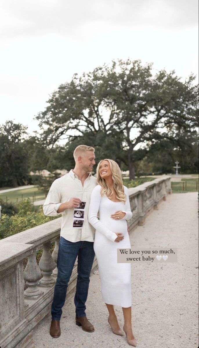 a pregnant couple standing on a bridge in front of some trees and bushes with their arms around each other