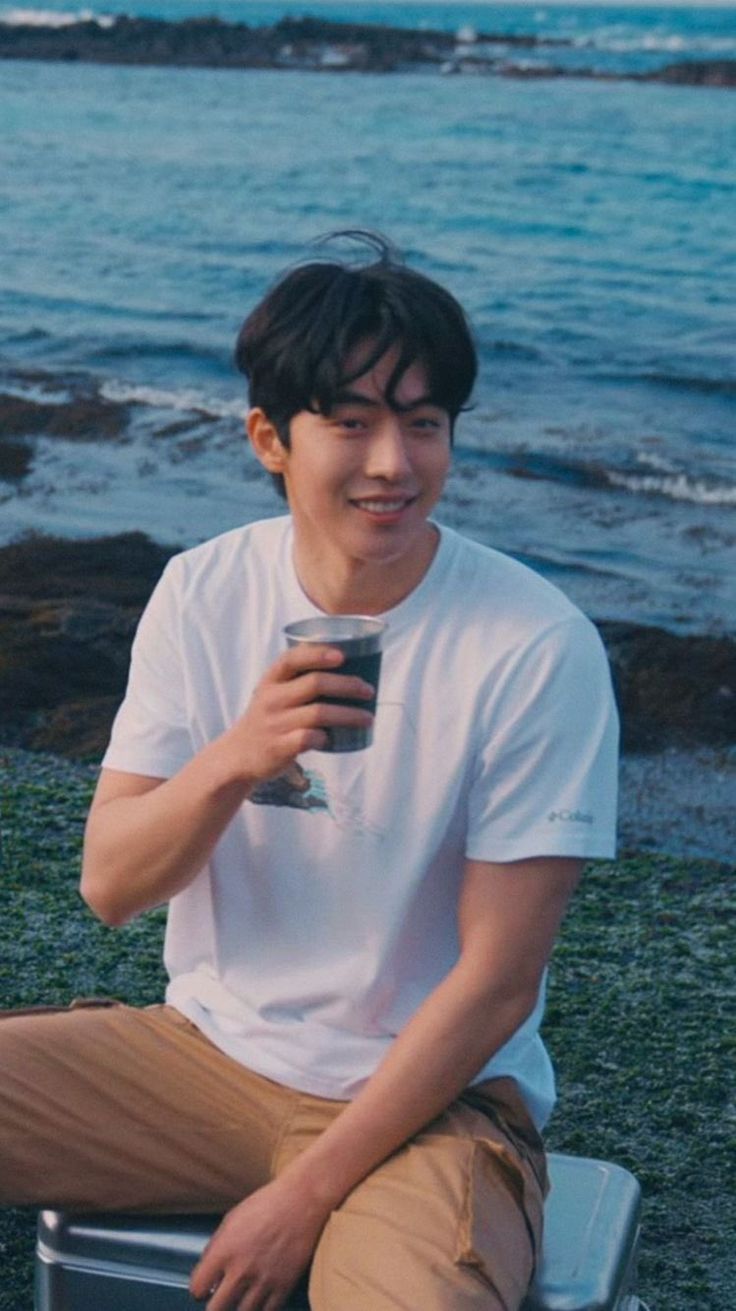 a young man sitting on top of a beach next to the ocean holding a drink