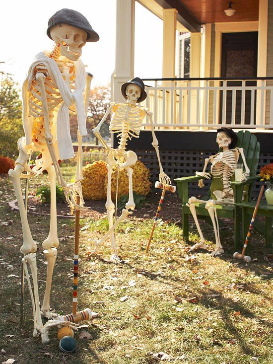 two skeleton statues in front of a house with lawn chairs and pumpkins on the ground