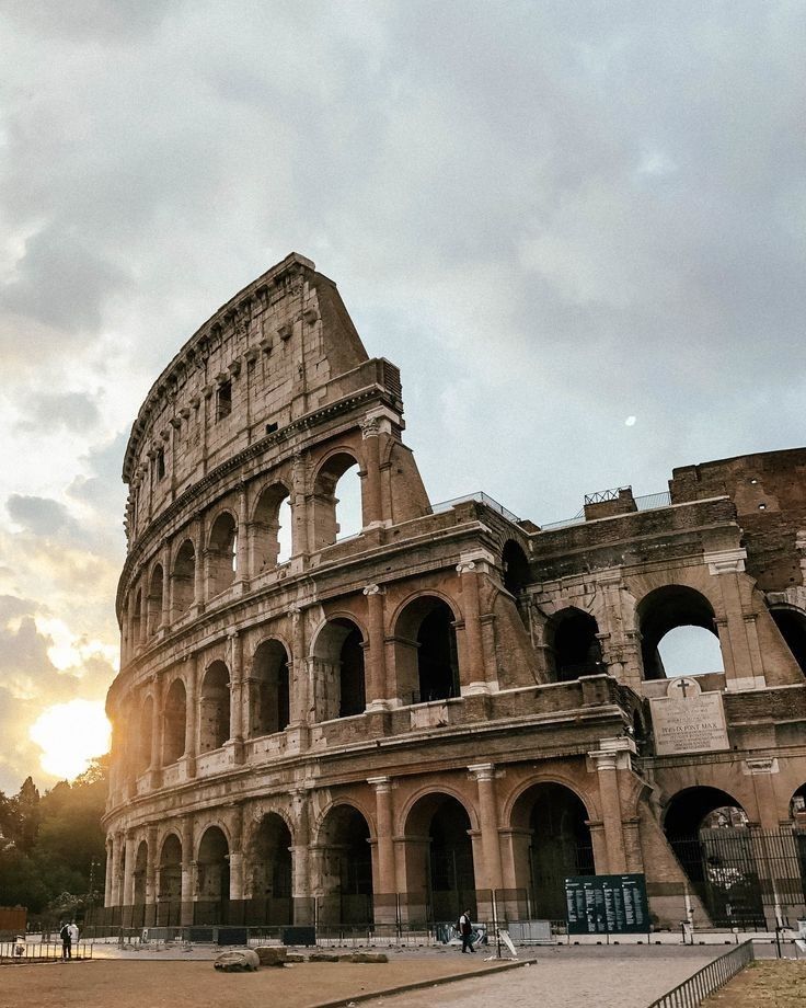 the sun is setting in front of an old roman colossion with people walking around it