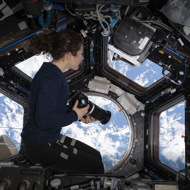 a woman holding a camera while looking out the window of a space station with earth in the background