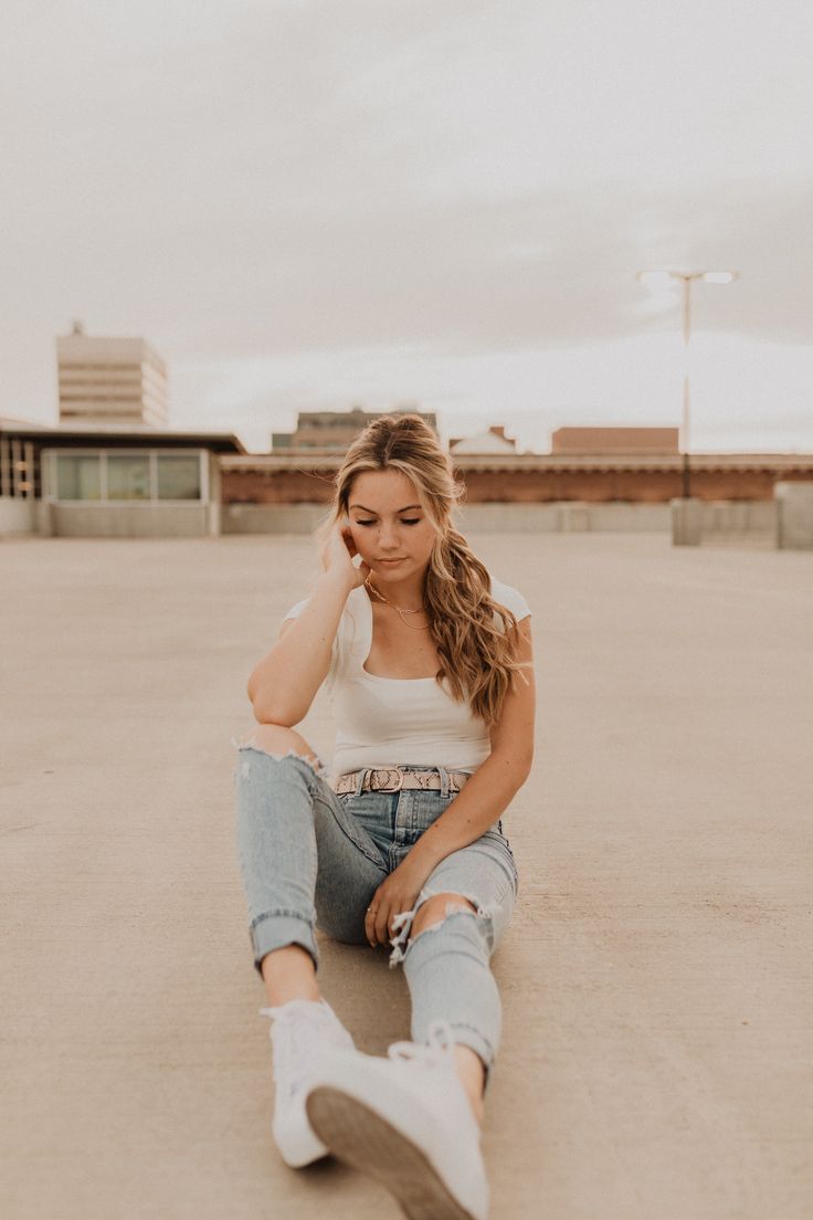 a woman sitting on the ground with her legs crossed, wearing ripped jeans and a white tank top