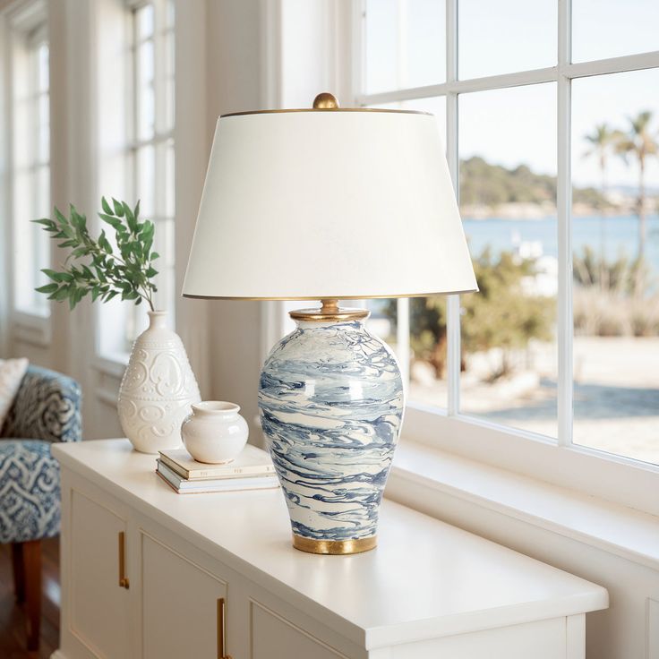 a blue and white vase sitting on top of a table next to a lamp in front of a window