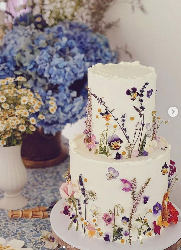 a three tiered cake decorated with flowers on a table next to blue and white hydrangeas