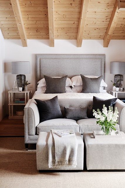 a bed sitting under a wooden ceiling next to a white chair and ottoman in front of a window