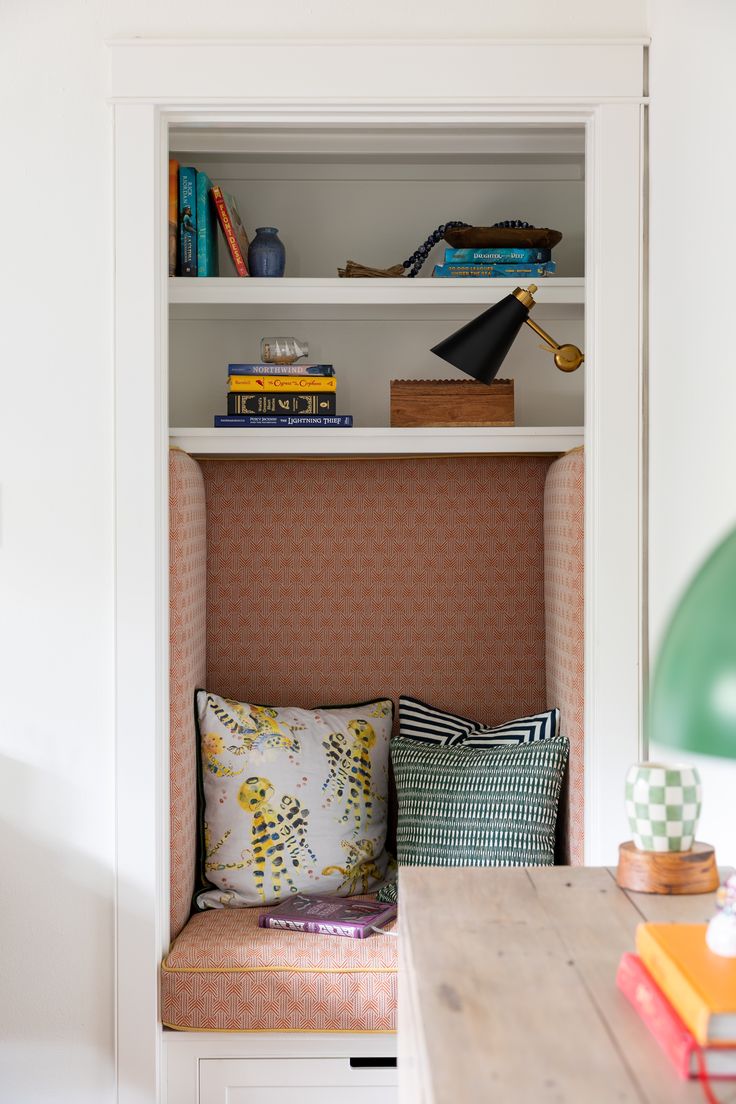 a room with some books and pillows on the shelf next to a table in front of it