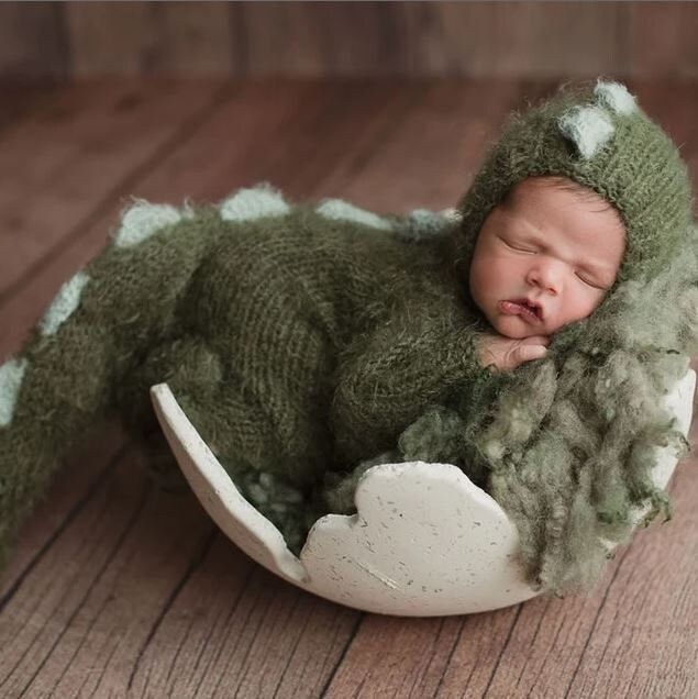 a newborn baby in a green knitted frog outfit sleeping on a white egg shell