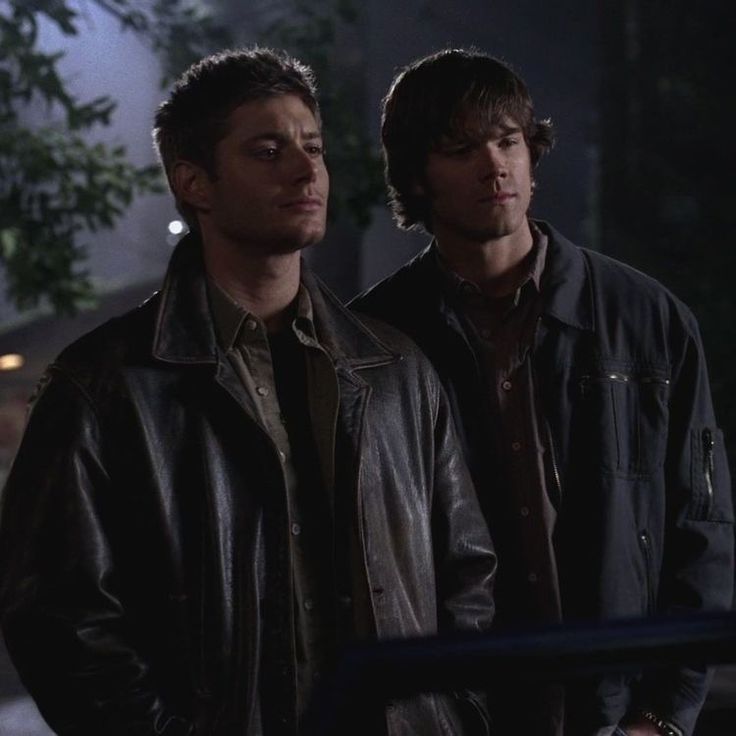 two young men standing next to each other in the street at night, one wearing a leather jacket