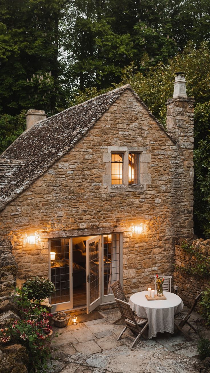 a small stone house with a table and chairs in front of it, surrounded by greenery