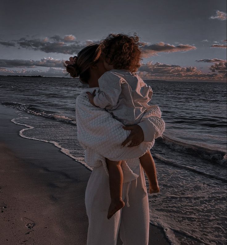 a woman holding a child on the beach at sunset with clouds in the sky behind her