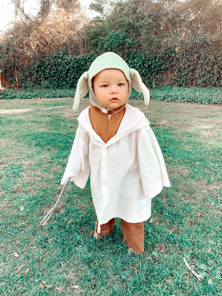 a baby dressed up in a costume standing on the grass with an animal behind him