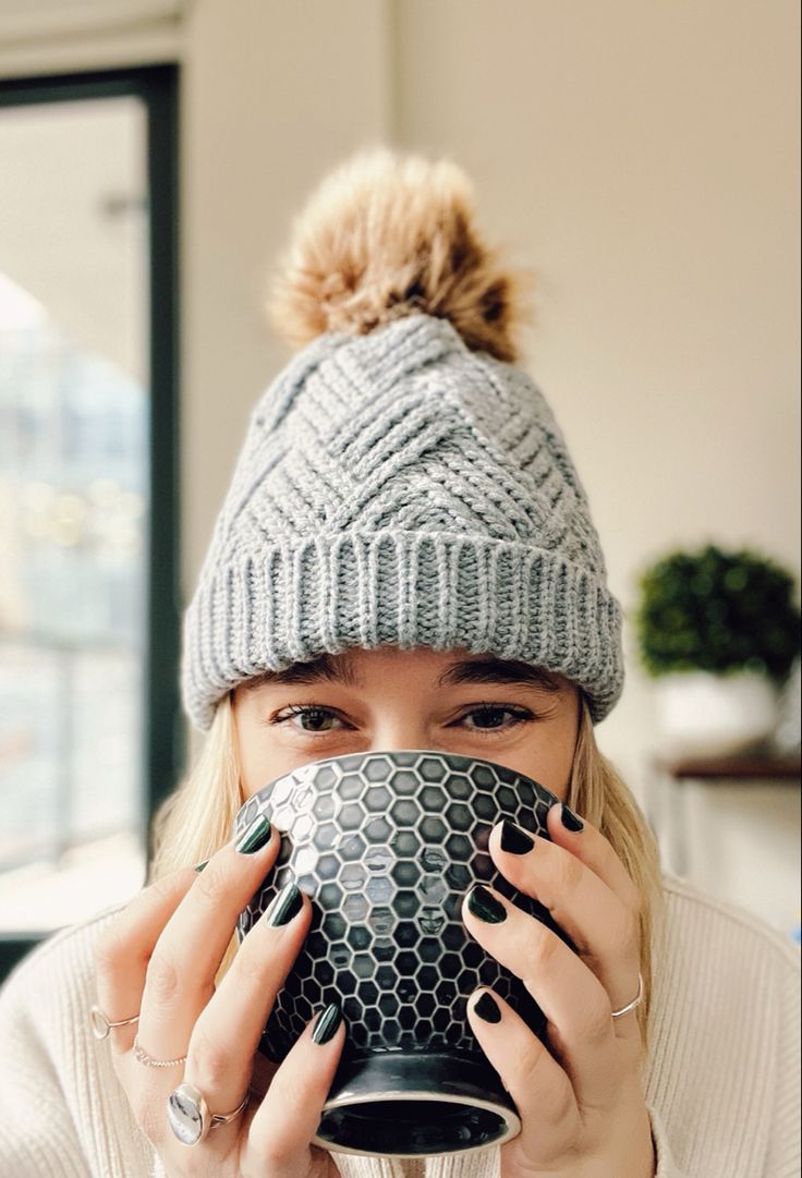 a woman holding a coffee cup in front of her face while wearing a knitted hat